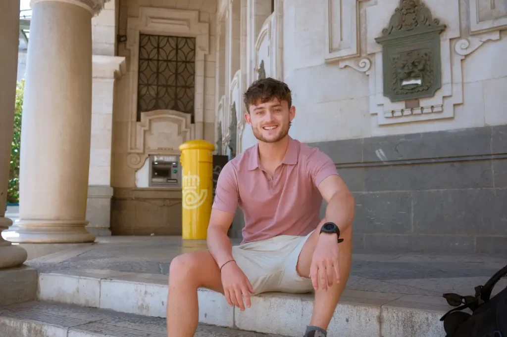 A picture of Garrett Landers, who  sits casually on stone steps outside an ornate historic building wearing pink polo shirt and beige shorts.