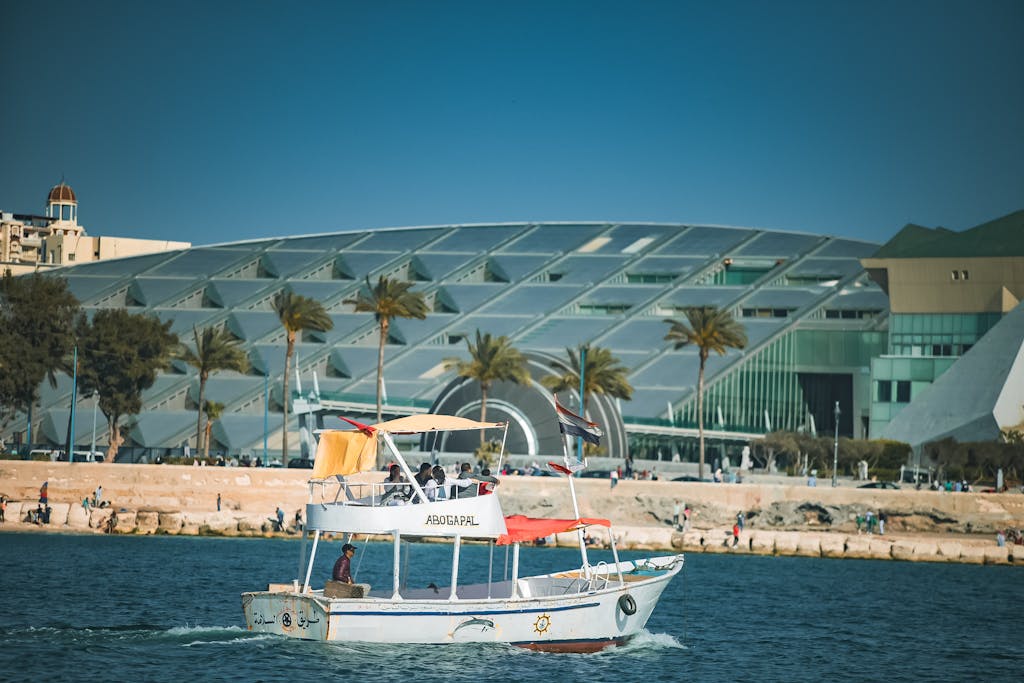 Boat in front of the modern Bibliotheca Alexandrina with palm trees and glass architecture.