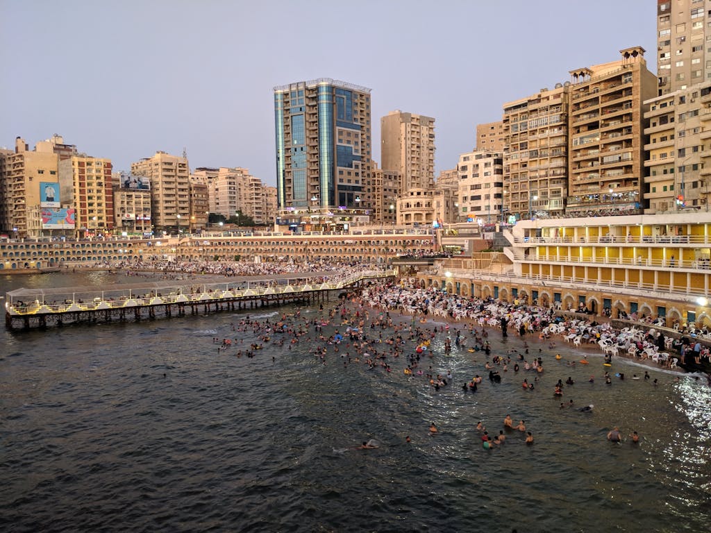 Crowded Alexandria public beach surrounded by high-rise buildings and a long pier.