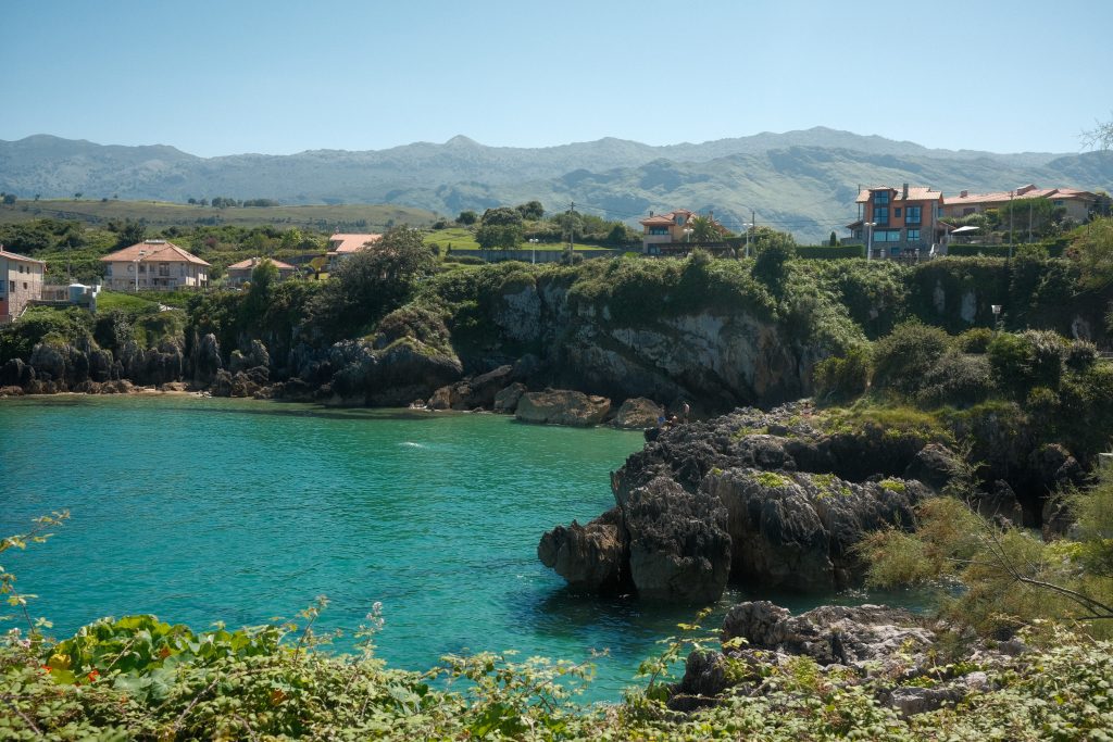 Coastal cliffs and turquoise waters in Llanes, Spain, with houses overlooking the sea and mountains in the background.