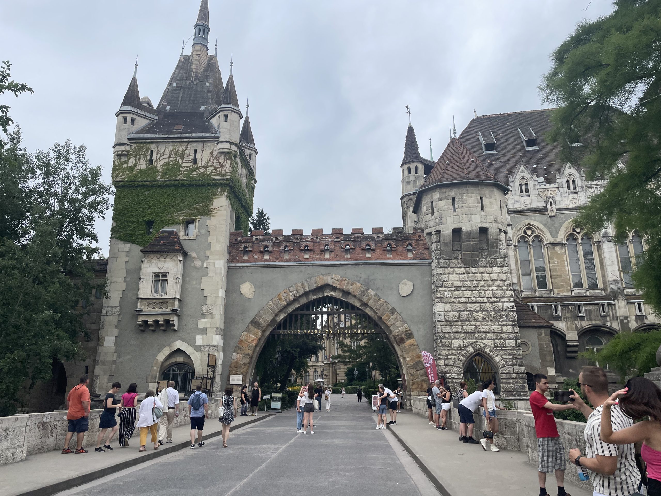 Entrance to Vajdahunyad Castle in Budapest with ivy-covered towers and tourists walking across the bridge