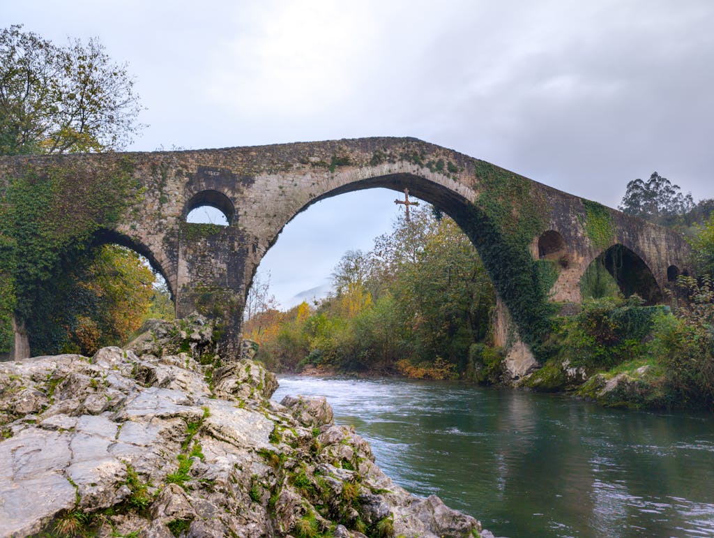 Scenic view of an ancient Roman stone bridge over the Sella River in Asturias, Spain, surrounded by lush autumn foliage.