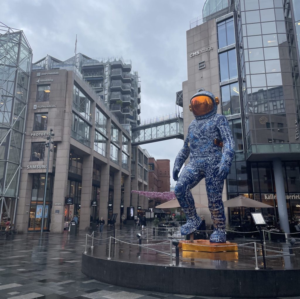 Colorful astronaut statue covered in graffiti-style writing at Aker Brygge in Oslo, Norway, surrounded by modern glass buildings on a rainy September afternoon.