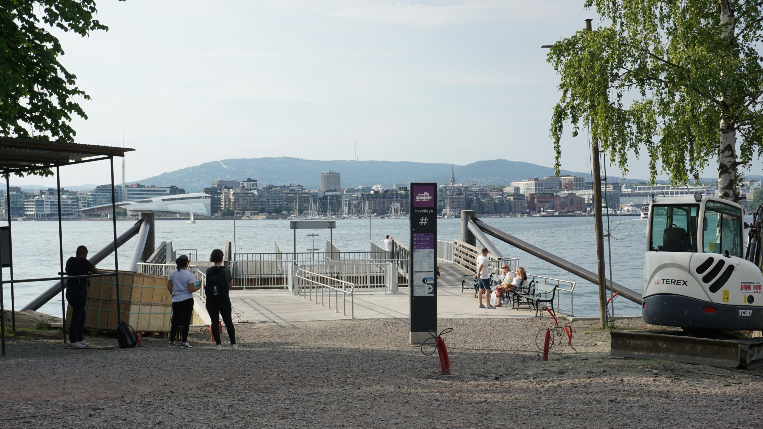 View of Oslo’s harbor from the Aker Brygge ferry terminal, with people sitting by the water and the city skyline visible across the fjord on a mild September afternoon.