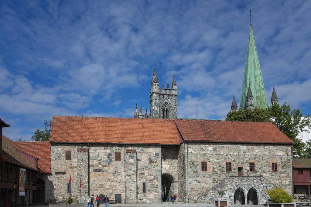 Exterior of the Archbishop’s Palace beside Nidaros Cathedral in Trondheim, Norway, with its red roof and medieval stone walls under a bright blue sky.