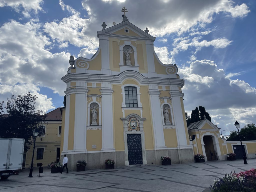 Carmelite Church in Győr, Hungary, with yellow and white baroque façade under dramatic clouds.