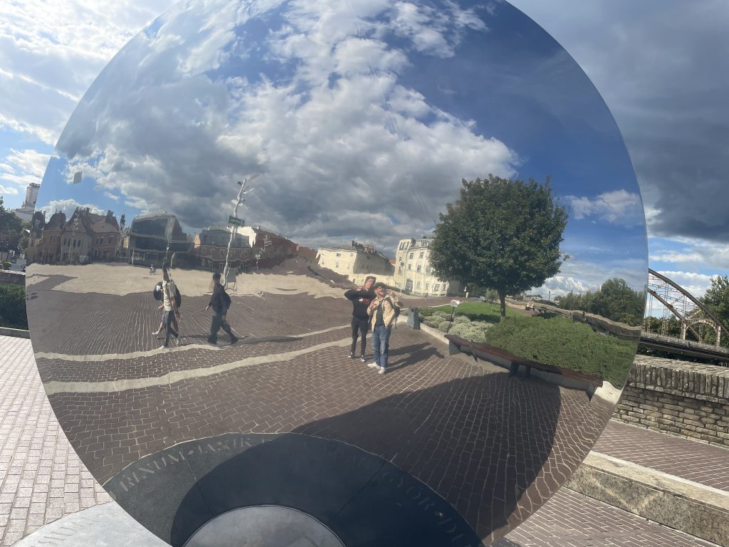 Reflective public art sculpture near Bécsi kapu Square in Győr, Hungary, showing the square and people walking by on a sunny afternoon.