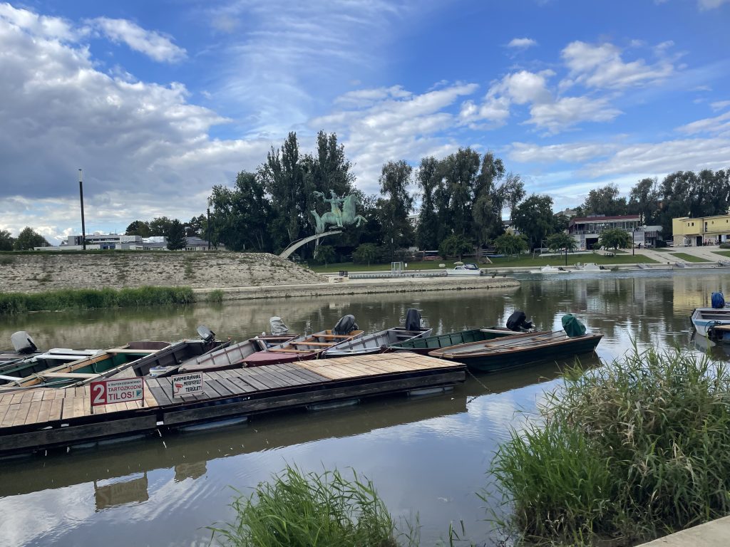 Small motorboats docked along the Rába River in Győr, Hungary, near the Danube confluence and the equestrian statue of Saint Stephen.