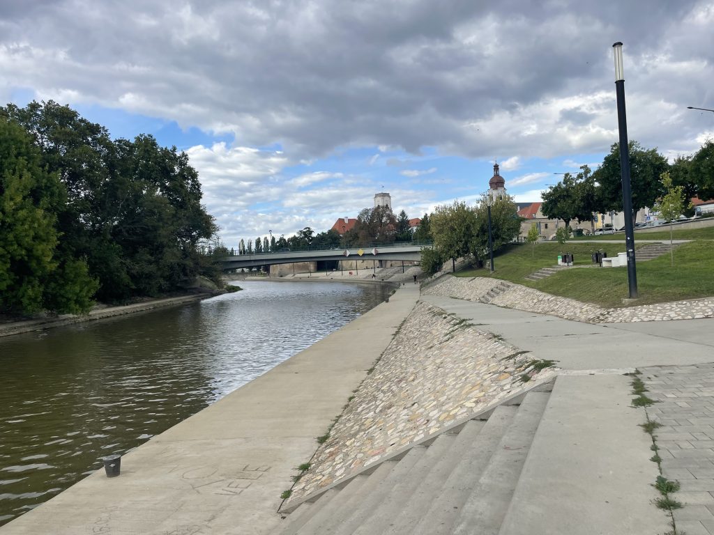 Walking path along the Rába River in Győr, Hungary, with a bridge and church towers visible in the distance.