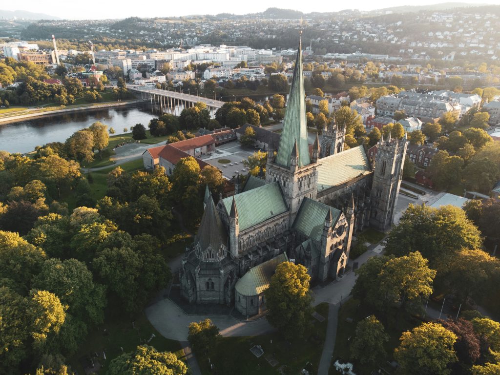 Aerial view of Nidaros Cathedral in Trondheim, Norway, surrounded by green trees, river, and cityscape. \