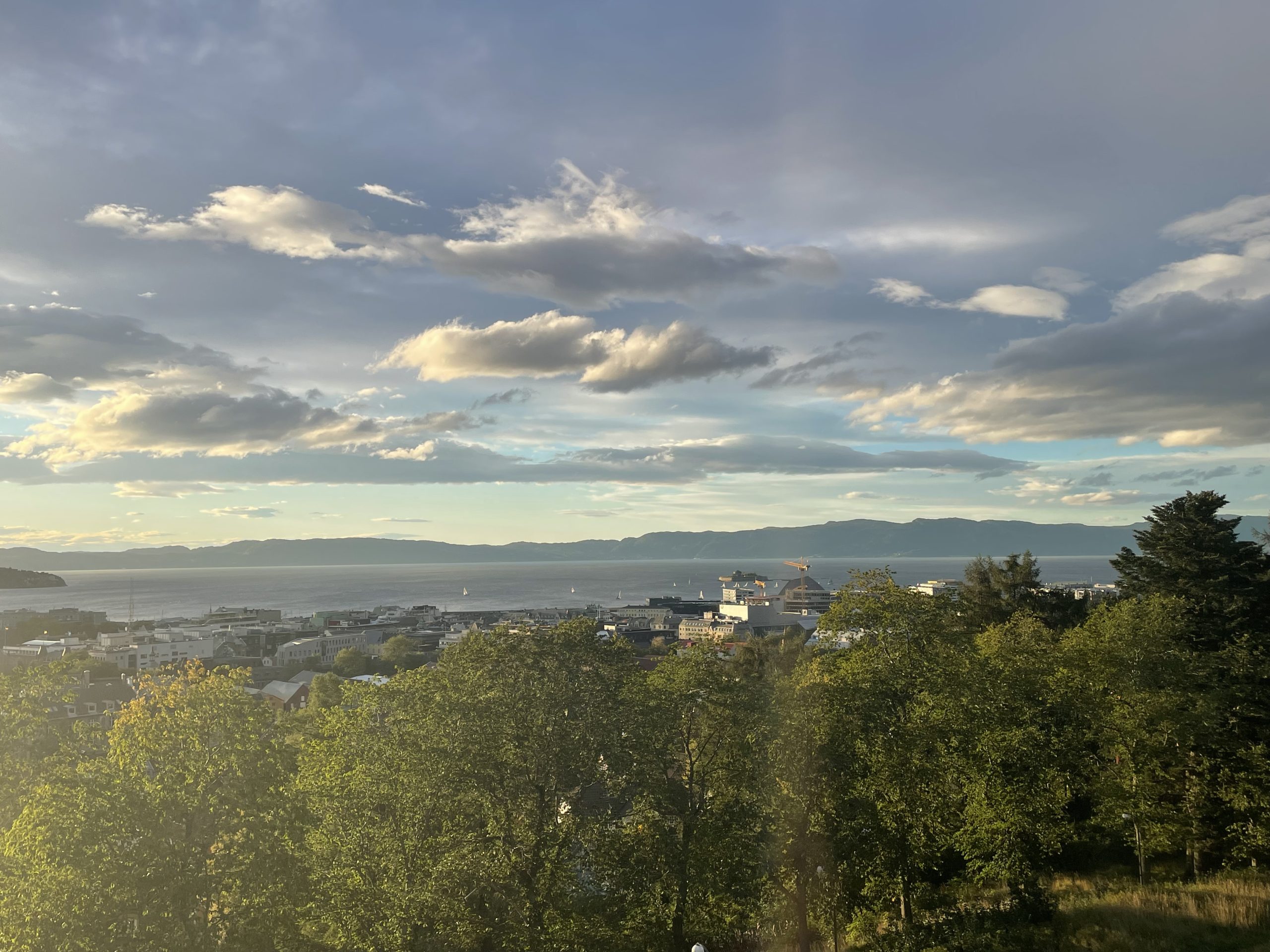 View over Trondheim, Norway from Kristiansten Fortress at sunset, showing the city, Trondheimsfjord, and surrounding mountains under a soft golden sky.