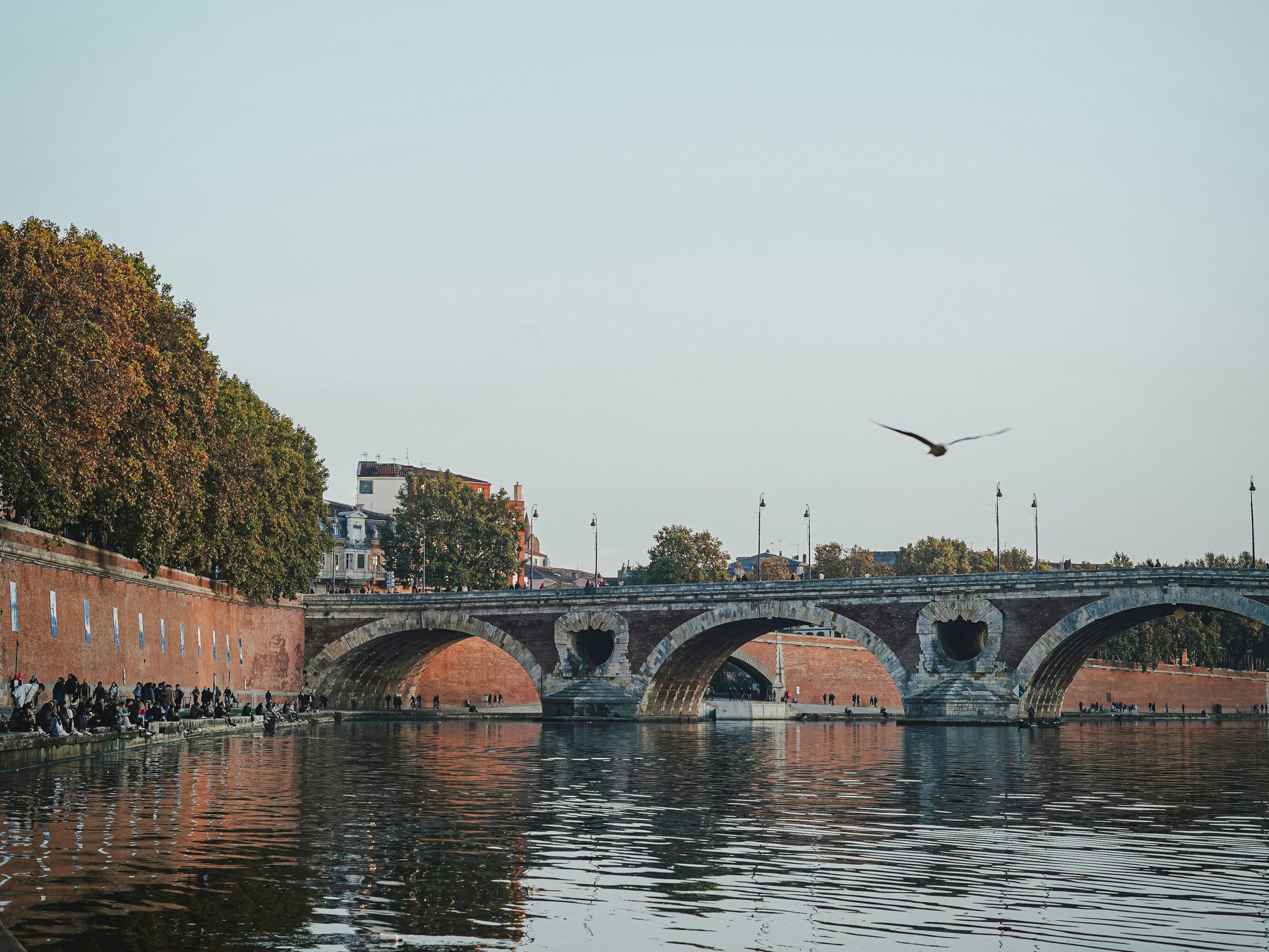 Pont Neuf and Garonne River in Toulouse at sunset with people sitting along the brick riverbank